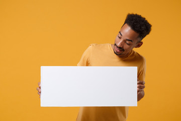 Curious young african american guy in t-shirt posing isolated on yellow orange background. People lifestyle concept. Mock up copy space. Hold white empty blank billboard with place for text or image.