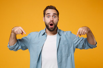 Shocked young bearded man in casual blue shirt posing isolated on yellow orange wall background in studio. People lifestyle concept. Mock up copy space. Pointing index fingers down keeping mouth open. - obrazy, fototapety, plakaty