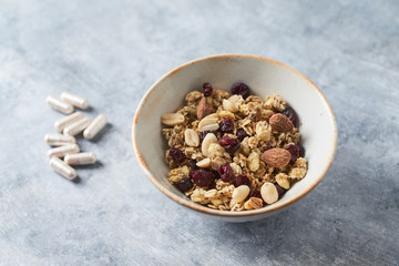 Bowl of granola with nuts,  cranberry, almonds and rasins. Sport supplements ( carnitine capsules ) in background. Bright stone  background. 