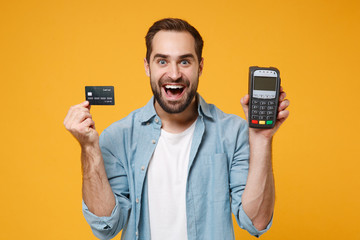 Excited young man in blue shirt posing isolated on yellow orange background. People lifestyle concept. Mock up copy space. Hold wireless bank payment terminal to process acquire credit card payments. - obrazy, fototapety, plakaty
