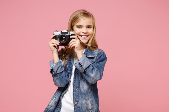 Smiling little blonde kid girl 12-13 years old in denim jacket posing isolated on pastel pink background in studio. Childhood lifestyle concept. Mock up copy space. Hold retro vintage photo camera.
