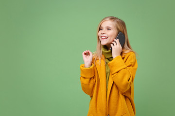 Smiling little blonde kid girl 12-13 years old in yellow coat posing isolated on pastel green background children portrait. Childhood lifestyle concept. Mock up copy space. Talking on mobile phone.