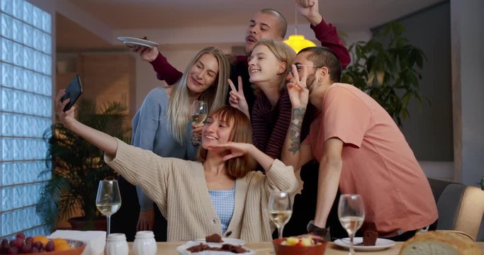 Happy friends making selfie together using black smartphone at house party celebration, cheerful smiling millennials holding mobile device to share front camera photo in social media indoor near table