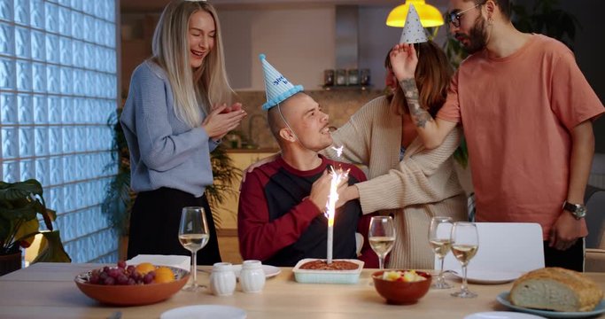 Happy Caucasian guy celebrating Birthday with friends and family together at home indoor, cheerful company clapping in hands around birthday boy in caps with tasty cake with candle fireworks on table