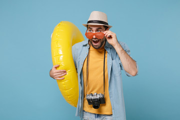 Surprised traveler tourist man in yellow clothes, eyeglasses with photo camera isolated on blue background. Passenger traveling abroad on weekends. Air flight journey concept. Hold inflatable ring.