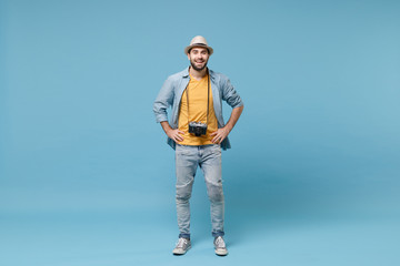 Smiling traveler tourist man in yellow summer clothes with photo camera isolated on blue background. Male passenger traveling abroad on weekends. Air flight journey. Stand with arms akimbo on waist.