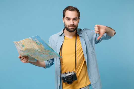 Dissatisfied traveler tourist man in casual clothes with photo camera isolated on blue background. Passenger traveling abroad on weekends. Air flight journey concept Hold city map showing thumb down.
