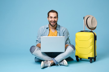 Cheerful traveler tourist man in summer casual clothes isolated on blue background. Passenger traveling abroad on weekend. Air flight journey concept. Sit near suitcase, work on laptop, booking hotel.