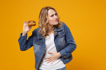 Dissatisfied young woman in denim clothes isolated on yellow orange background studio portrait. Proper nutrition or American classic fast food concept Hold burger put hand on stomach having ache pain.