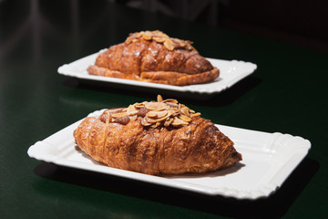 Delicious fresh baked almond croissants with filling on the white plate at the dark cafe. Green table. Focus on the foreground.