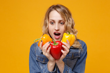 Shocked young woman in denim clothes isolated on yellow orange background in studio. Proper nutrition vegetarian food healthy lifestyle diet meal. Diabetes dieting concept. Hold colorful bell peppers.