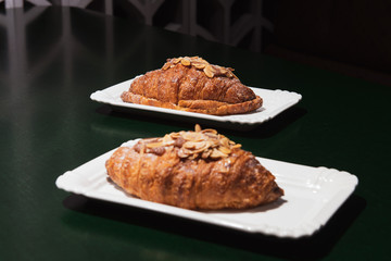 Delicious fresh baked almond croissants with filling on the white plate at the dark cafe. Green table. Focus on the background.