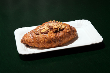 Delicious fresh baked almond croissant with filling on the white plate at the dark cafe. Green table. Closeup.