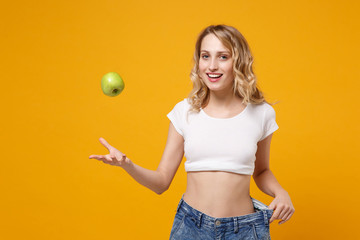 Smiling young woman in white t-shirt isolated on orange background. Proper nutrition losing weight healthy lifestyle dieting concept. Wearing old big jeans showing her diet results throwing up apple.