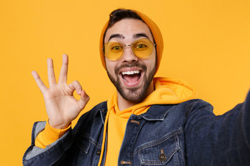 Close up of hipster guy in fashion jeans denim clothes posing isolated on yellow orange background. People lifestyle concept. Mock up copy space. Doing selfie shot on mobile phone, showing OK gesture.
