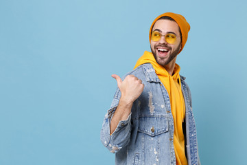 Side view of laughing young hipster guy in fashion jeans denim clothes posing isolated on pastel blue background studio portrait. People lifestyle concept. Mock up copy space. Pointing thumb aside.