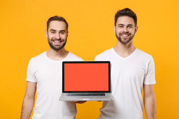 Smiling young men guys friends in white t-shirts posing isolated on yellow orange background in studio. People lifestyle concept. Mock up copy space. Hold laptop pc computer with blank empty screen.