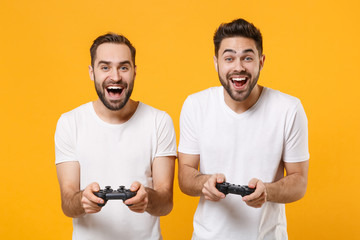 Fototapeta na wymiar Laughing young men guys friends in white t-shirts posing isolated on yellow orange background studio portrait. People sincere emotions lifestyle concept. Mock up copy space. Play game with joystick.