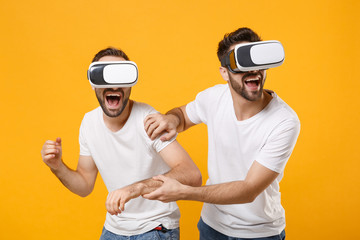 Cheerful young men guys friends in white blank empty t-shirts posing isolated on yellow orange background studio portrait. People emotions lifestyle concept. Mock up copy space. Watching in headset.