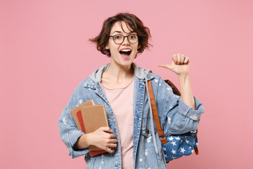 Naklejka premium Excited young woman student in denim clothes eyeglasses backpack posing isolated on pastel pink background. Education in high school university college concept. Hold books pointing thumb on herself.