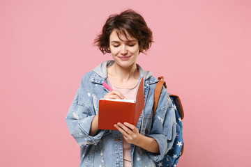 Smiling young woman student in denim clothes backpack isolated on pastel pink background in studio. Education in high school university college concept. Mock up copy space. Writing notes in notebook.
