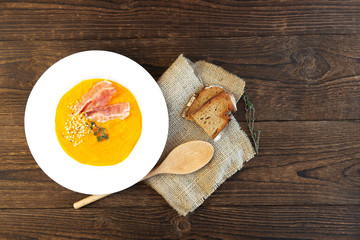 Autumn pumpkin soup on a rustic old wooden table with slices of bacon and hemp seeds in a white plate. Nearby lies a wooden spoon and slices of bread. Place for text
