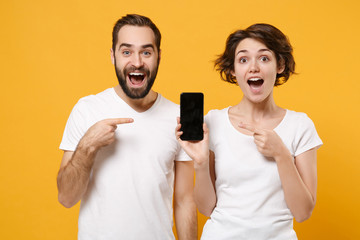 Amazed couple friends bearded guy girl in white t-shirts isolated on yellow orange background. People lifestyle concept. Mock up copy space. Point index finger on mobile phone with blank empty screen.