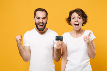 Excited couple friends bearded guy girl in white t-shirts isolated on yellow orange background. People lifestyle concept. Mock up copy space. Hold credit card, doing winner gesture, showing thumb up.