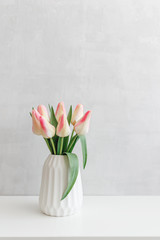 Light pink tulips in a white geometric ceramic vase stand on a white table near grey wall.