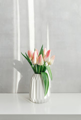 Light pink tulips in a white geometric ceramic vase stand on a white table near grey wall. Bouquet of flowers in the morning sun beams.