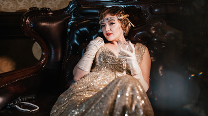 a girl in a gold dress with a diadem on her head, sits on a leather armchair, next to a gun and...