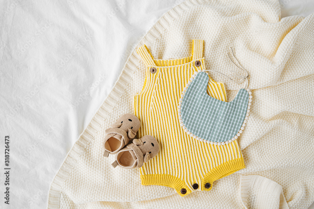 Wall mural Yellow bodysuit, bib, baby boots and toy on knitted blanket. Set of  kids clothes and accessories  on bed. Fashion newborn. Flat lay, top view