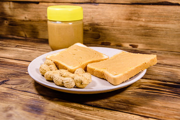 Glass jar with peanut butter and plate with sandwiches on a wooden table
