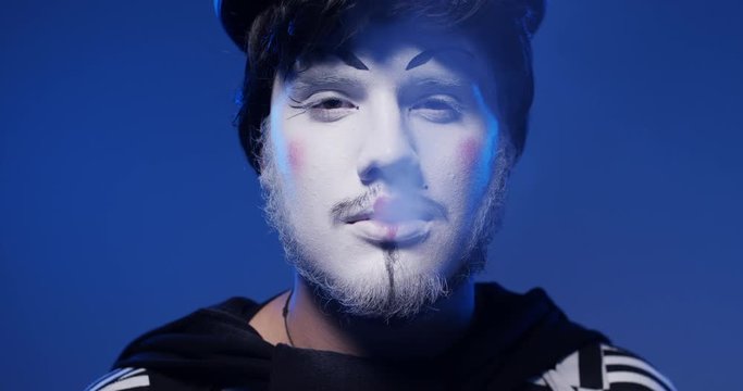 Close up of handsome male mime smiling into a camera and smoking cigarette in black and white striped clothes. Isolated blue background