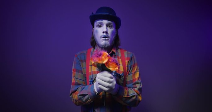 Handsome colorful charismatic mime clown in a hat making trembling, nervous emotions with a bouquet of flowers in his hands, looks uncertainly at the camera