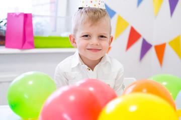 Fototapeta na wymiar Portrait of blonde caucasian boy smiling at camera near birthday rainbow cake. Festive colorful background with balloons.
