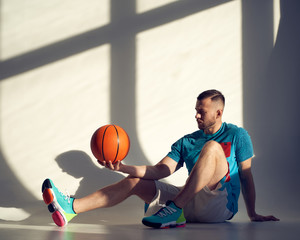 Young athletic man, basketball player holding ball on shoulder and sitting near wall with shadows...