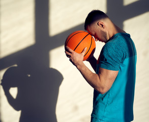 Young athletic man, basketball player holding ball and standing near wall with shadows from window