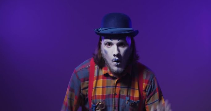 Handsome colorful charismatic mime clown in a hat dances emotionally on purple isolated background.