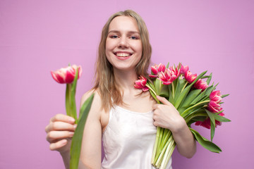 attractive girl holds a bouquet of flowers on a pink background and gives one tulip, the seller offers flowers, a woman gives tulips