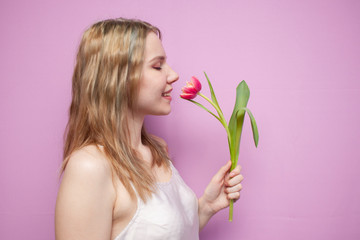 Obraz na płótnie Canvas young attractive girl with one tulip on a colored pink background, a woman holds a flower and sniffs