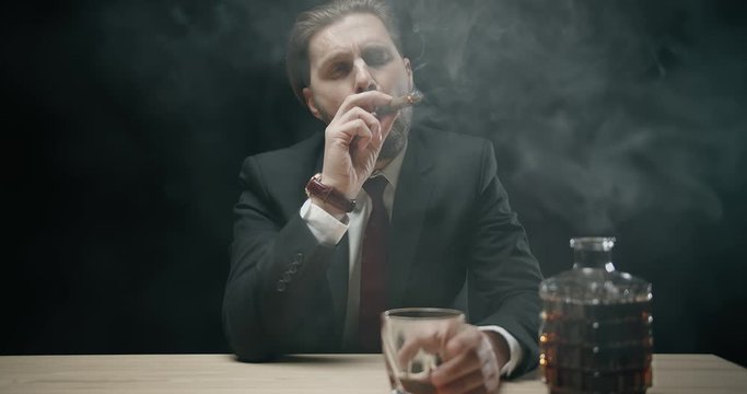 Brutal businessman in elegant suit drinking whiskey and smoking cigar isolated on black background