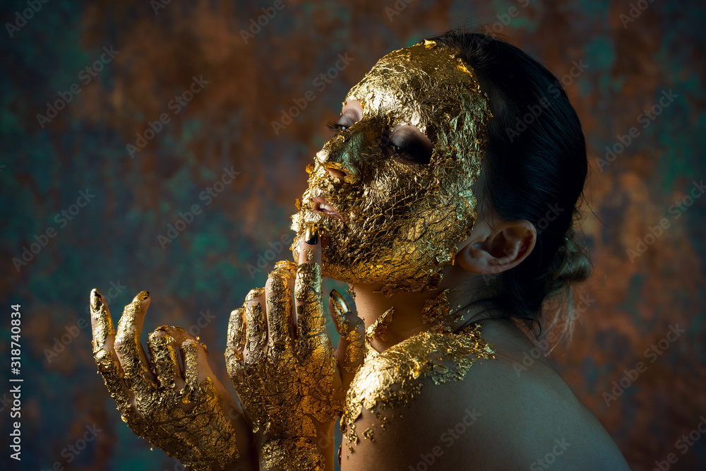 Wall mural Girl with a mask on her face made of gold leaf. Gloomy studio portrait of a brunette on an abstract background.