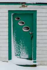 A white building made of wood clapboard with a green door covered in snow. The step is covered in snow too. The vintage building is textured and worn. The door has three small oval windows and a lock.