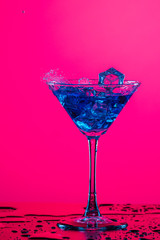 Alcohol Drink Spilling Out of The High Glass Goblet with Ice Cubes and Colorful Liquid. Isolated Over Colorful Background.