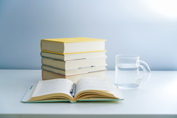 An open book is placed on a white desktop. A stack of neatly arranged books and a transparent glass on a white table