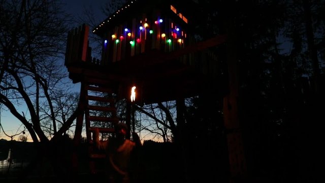 A young boy standing on an illuminated treehouse at night holding a torch