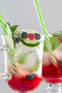 Homemade cold summer cocktail with mineral water, ice cubes, mint, lime, berry juice and fresh berries in glasses with straws, vertical image