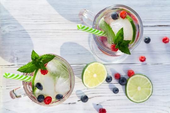 Homemade cold summer cocktail with mineral water, ice cubes, mint, lime, berry juice and fresh berries in glasses with straws, top view