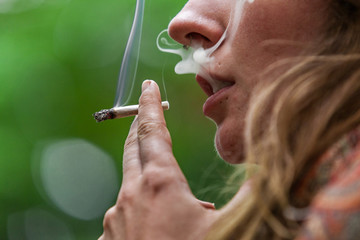 Low angle closeup cropped shot of caucasian woman's face with hand rolled marijuana joint. Smoke escaping from girl's nose and mouth outside.